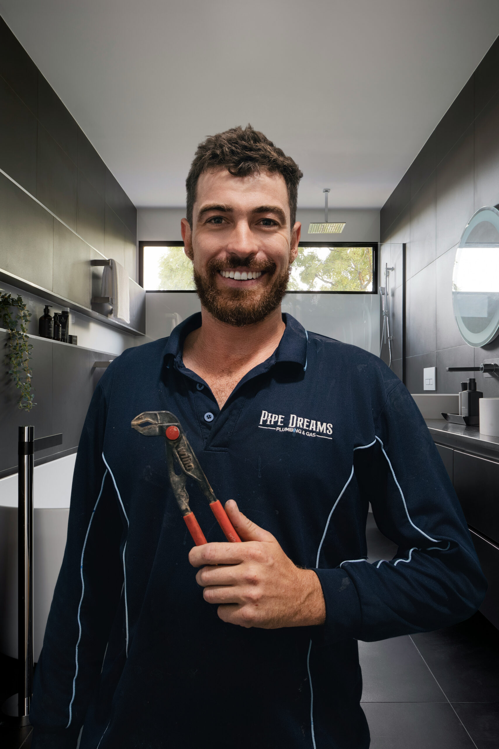 Handsome plumber smiling and holding multi-grips tool in work uniform standing front on against a freshly renovated bathroom photoshoot for About Us page